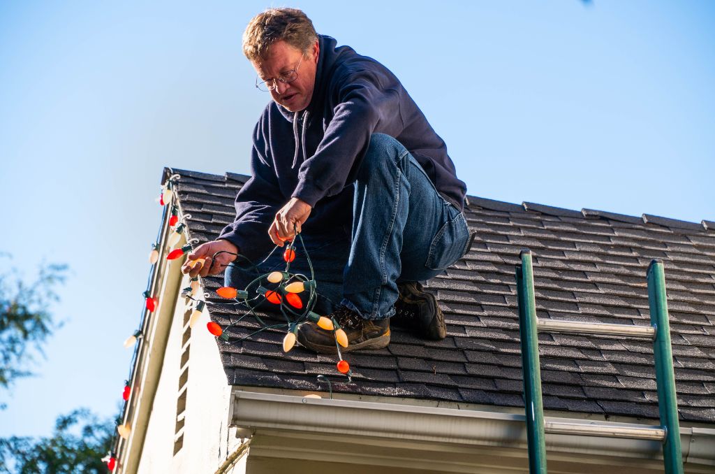 Man on his roof installing Christmas lights with a ladder in the foreground.
